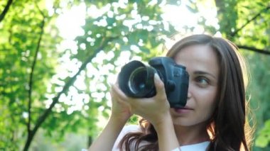 Beyaz gömlek giyen Oldukça gülümseyen kız fotoğrafçı yeşil yaprakları yumuşak bir arka plan üzerinde bir parkta fotoğraf yapıyor. Bir kadın gün batımında güneşin ışınlarına bakarak doğayı fotoğraflar..