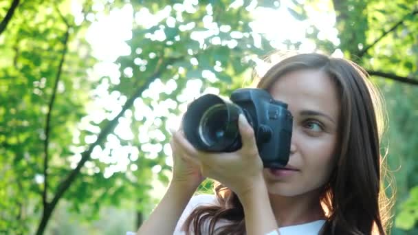 Una fotógrafa muy sonriente con una camisa blanca está haciendo fotos en un parque sobre un fondo suave de follaje verde. Una mujer fotografía la naturaleza mirando a través de los rayos del sol al atardecer . — Vídeo de stock