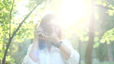Beyaz gömlek li oldukça gülümseyen kız fotoğrafçı yeşil yaprakları yumuşak bir arka plan üzerinde bir parkta fotoğraf yapıyor. Bir kadın gün batımında güneş ışınları ile kameraya bakarak model fotoğrafları.