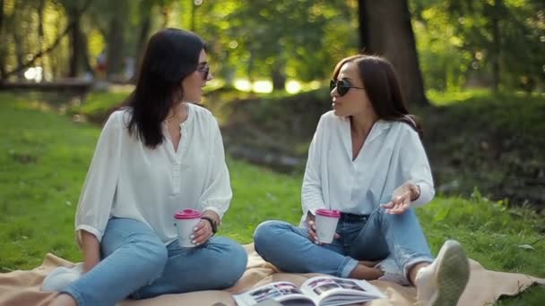 Dos elegantes chicas sonrientes con camisas blancas se sientan y se ríen en el parque sobre un fondo suave de follaje verde. Las novias se divierten, hablan, beben café en tazas de papel y se registran . — Vídeo de stock