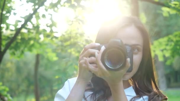 Güzel gülümseyen ve beyaz gömlek giyen gülen kız fotoğrafçı yeşil yaprakları yumuşak bir arka plan üzerinde bir parkta fotoğraf yapıyor. Bir kadın gün batımında güneş ışınları ile doğa fotoğrafları. — Stok video