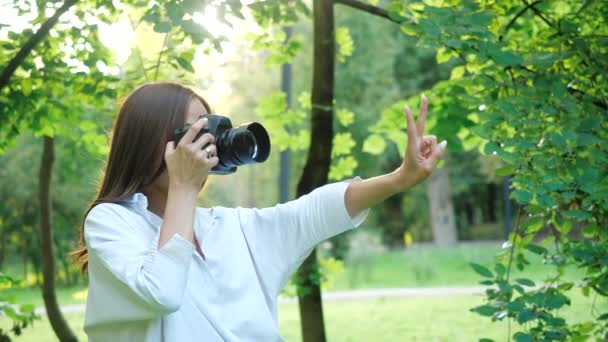 Krásná holka fotografka v bílé košili dělá fotky a než si vyfotí fotku, tak se domnívá, že má prsty: jedna, dvě, tři. Žena fotografovat skrz paprsky slunce. — Stock video