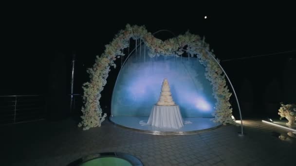 Mooie smakelijke bruidstaart onder versierde boog tegen blauwe achtergrond 's nachts. Steadicam schot — Stockvideo