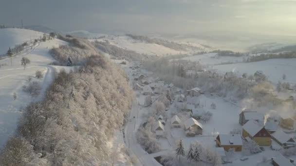 Lot nad wioską w Karpatach zimą o wschodzie słońca. Widok z lotu ptaka na ośnieżone domy w górach. Krajobraz wiejski zimą. Górska wioska w śniegu z wysokości. — Wideo stockowe
