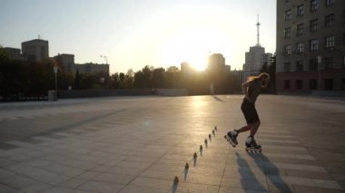 Genç uzun saçlı adam patenci gün batımında bir şehir parkında akşam konileri arasında dans ediyor. Serbest stil slalom Pateni Konileri arasında.