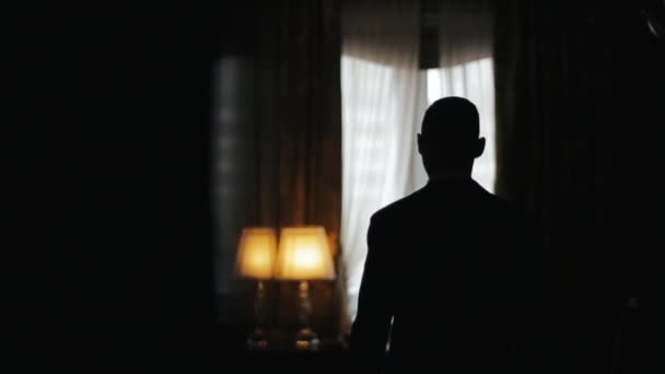 Silhouette d'un homme portant un costume marchant d'une pièce sombre vers une fenêtre lumineuse au ralenti. L'homme s'arrête, met les mains sur une table et semble cher à la fenêtre . — Video