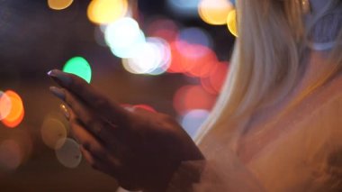 Şehirde gece vakti akıllı telefonlu sarışın kadın ellerinin yakın çekimi. Geceleri şehir caddesinde akıllı telefondan mesaj yazmak. Akıllı telefondan mesaj atan kız.