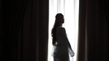 Güzel, seksi bir kızın silueti pencereden yansıyan sıcak bir sabah ışığına karşı poz veriyor. Güzel kız sabahleyin pencere perdesini açar. Kameranın önünde seksi yumuşak hareketler.