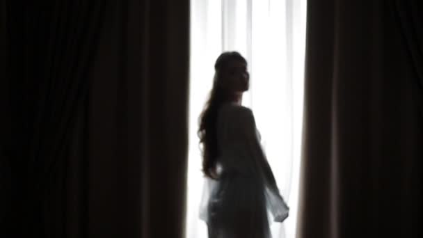 Silhueta de uma menina sexy bonita posando contra uma luz da manhã quente brilhando pela janela. Menina bonita abre cortina de janela pela manhã. Movimentos suaves Sexy na frente da câmera . — Vídeo de Stock