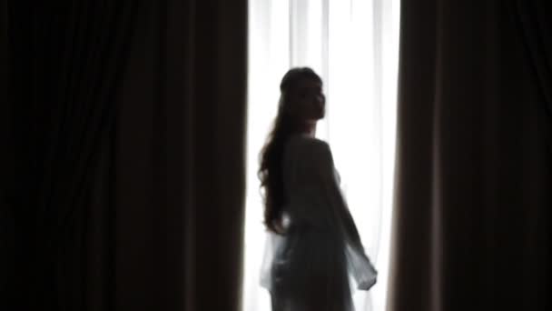 Silhouette di una bella ragazza sexy in posa contro una calda luce del mattino che brilla attraverso la finestra. Bella ragazza apre la tenda della finestra al mattino. Movimenti morbidi sexy davanti alla fotocamera . — Video Stock