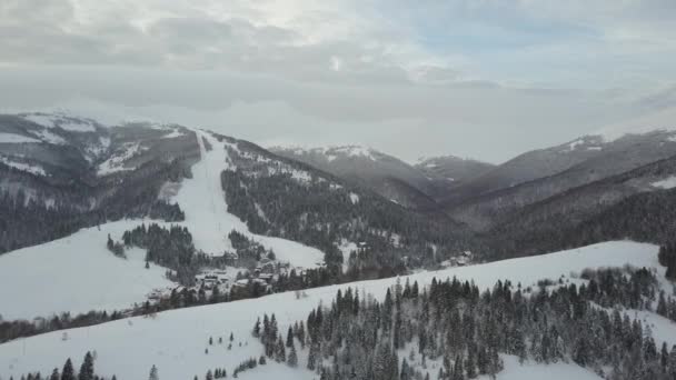 カルパチア山脈の村とそれに隣接するスキーリゾートの上を飛行します。山の雪に覆われた家々の鳥の視点。冬の田舎の風景。高さから雪の中のカルパチアの村. — ストック動画