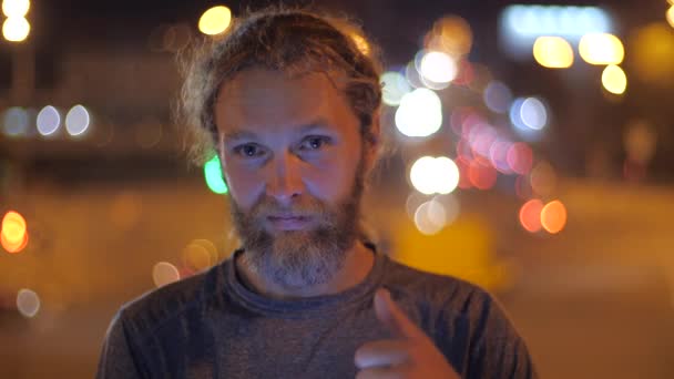 笑顔ハンサムな白人ひげを生やした長髪の若い男はカメラにまっすぐに見え、都市の移動する夜の光の背景に親指を表示します. — ストック動画