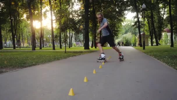 Freestyle Rolschaatsen bij zonsondergang. Jonge langharige man met baard rolschaatser danst tussen kegels in een leuke avond in een stadspark met duim omhoog. — Stockvideo