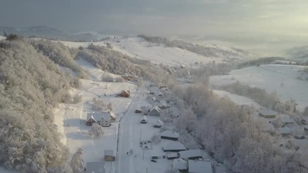 Górska wioska w śniegu z wysokości. Lot nad wioską w Karpatach zimą o wschodzie słońca. Widok z lotu ptaka na ośnieżone domy w górach. Krajobraz wiejski zimą. — Wideo stockowe