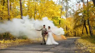 Ağır çekim. Genç ve çekici gelin ve damat yanan sis bombaları tutuyor ve sonbahar ormanlarında yol boyunca yürüyorlar. Parktaki renkli sonbahar ağaçları arasında mutlu yeni evliler..