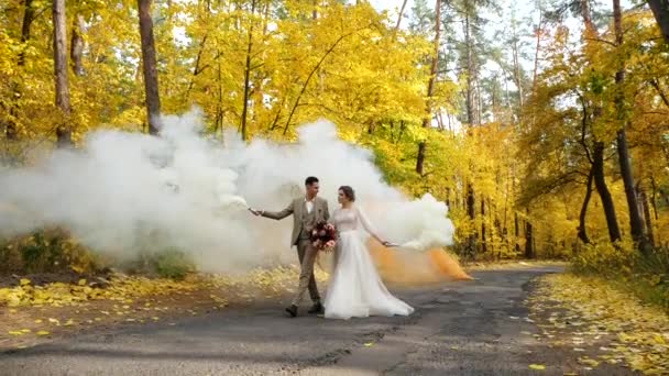 Movimento lento. Jovem e atraente noiva e noivo segurar queima bombas de fumaça e caminhar ao longo de uma estrada na floresta de outono. Feliz amor recém-casados entre árvores de queda coloridas em um parque . — Vídeo de Stock