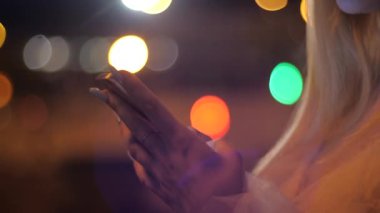 Şehirde gece vakti akıllı telefonlu sarışın kadın ellerinin yakın çekimi. Geceleri şehir caddesinde akıllı telefondan mesaj yazmak. Akıllı telefondan mesaj atan kız.