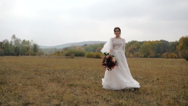 カラフルな森と美しい自然景観に対する幻想的な秋の牧草地でスローモーションで結婚式の花束と陽気な花嫁が回る. — ストック動画
