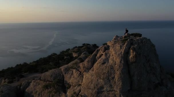 Vzdušná silueta mladé úspěšné ženy stojící na vrcholu hory proti moři při východu slunce. Žena horolezec na vrcholu hory na Krymu s nataženými pažemi. — Stock video