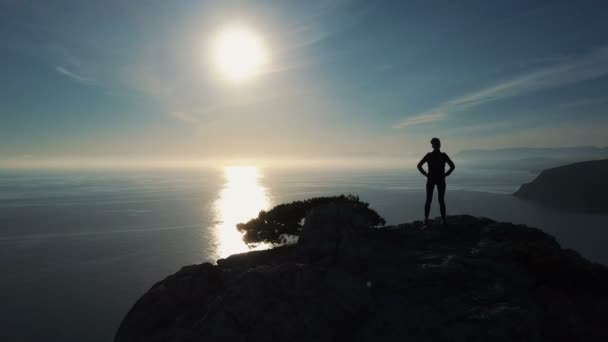 Silhouette aérienne de la jeune femme confiante qui réussit alpiniste sur le sommet de la montagne en Crimée avec les bras tendus. Femme courageuse debout sur le sommet d'une montagne contre la mer au coucher du soleil . — Video
