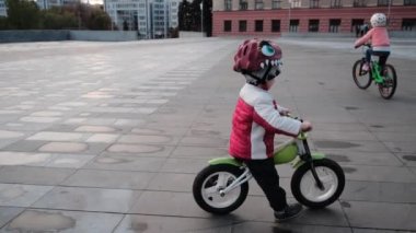 Küçük gülümseyen mutlu çocuk ve kız şehir parkında bisiklet sürüyorlar, yakın plan. Kask takan küçük motorcu şehir parkında bisiklet sürüyor..