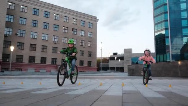 Little smiling happy girl and boy ride green bicycles by a circle in evening in a city. Little Bikers with helmets follow a moving camera. — Stock Video