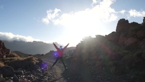 Вид сзади на прогулочную туристку в розовой куртке на каменной горной дороге в солнечный весенний день на острове Тенерифе на фоне потрясающего горного пейзажа, покрытого облаками . — стоковое видео