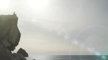 Kıyıya yakın bir uçurumun kenarında oturan bir adamın altından ateş etmek. Bir turistin silueti taştan bir uçurumun kenarına çömelmiş ve deniz ve güneşin güzel manzarasına hayran kalmıştır..