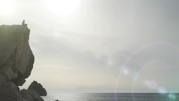 海岸近くの崖の端に座っている男の下から撮影。石の崖に囲まれた観光客のシルエットと海と太陽のまぶしさの美しい自然の景色を賞賛. — ストック動画