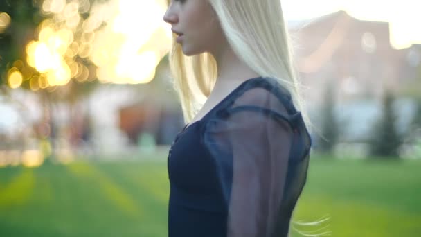 Filmado de cerca cinematográfico en el perfil de una impresionante joven rubia europea con el pelo largo en negro caminando en el parque de la ciudad y sonriendo a la cámara, el viento efectivamente sopla su cabello sedoso en cámara lenta — Vídeos de Stock