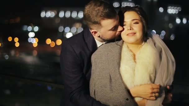 O par de casamento jovem feliz beija-se em uma varanda de restaurante de tarde em um contexto embaçado de lanternas e tráfego. Groom em um terno beija sua noiva, protegido por uma jaqueta fora no terraço . — Vídeo de Stock