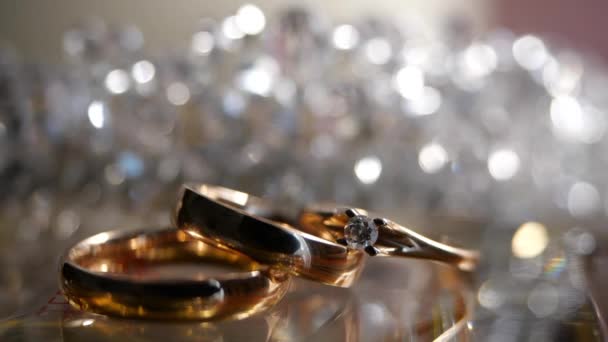 Három gyönyörű arany gyűrű, egy gyémánt eljegyzési gyűrű és egy eljegyzési gyűrű áll csillogó és csillogó háttérrel szemben. Ajándékok és romantika, dekoráció, menyasszony és vőlegény, irizáló fény és csillogás — Stock videók
