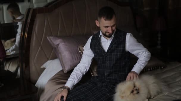Beau jeune homme barbu dans un costume élégant assis sur un lit luxueux dans un hôtel le matin et caresse son chien bien-aimé. Le gentil marié sourit et caresse le spitz sur le lit dans la chambre . — Video