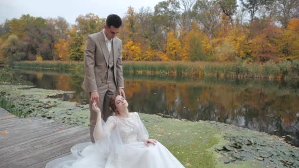 ウェディングドレスの美しい若い花嫁は目を閉じて木製の橋の上に座っており、秋の森を背景に小さな湖の近くの彼女の新郎の足にもたれています。新婚カップルをリラックスさせる. — ストック動画