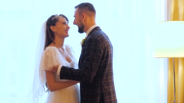 Feliz boda pareja sonrisas y abrazos en una acogedora habitación de un hotel de lujo en Kharkov, Ucrania. novio barbudo besa a su hermosa y querida novia en un vestido de novia cerca de las cortinas de la lámpara y la ventana . — Vídeo de stock