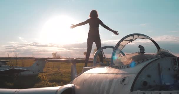 Задний вид человека, стоящего на советском военном истребителе времён Второй мировой войны на аэродроме и поднимающего руки вверх от удовольствия, смотрящего на большое солнце в замедленной съемке. Туристическая прогулка на кладбище самолетов . — стоковое видео