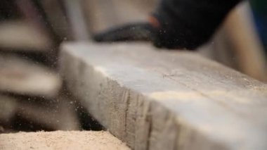 Adam öğütücüyle testereyi kestikten sonra tahtadaki talaşları uçuruyor ve onları eldivenli elle eziyor, yakın görüş. Dairesel bir makine kullanarak ahşap üzerinde çalıştıktan sonra havada talaş uçuşuyor..