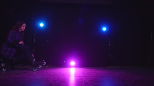 Naderend zicht op roze spotlicht tussen blauwe lampen en een rolschaatsend meisje zittend op voor- en achterwielen op donker podium. Concept van sport en gezonde levensstijl. Gieten in de studio. — Stockvideo
