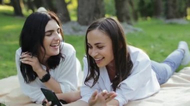 Beyaz tişörtlü ve kot pantolonlu güzel genç kız arkadaşların yakından görüntüsü çimlerin üzerinde uzanmış, akıllı telefona bakıyor ve güneşli yaz parkında şarkı söylüyor. Şirin kızlar doğada telefonla karaokeyi severler..