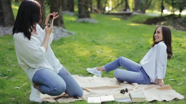 A lány fotózza a barátnőjét a városi napfényes parkban tavasszal okostelefonon. Két mosolygó barátnő rendezett fotózást a füvön ülve a fák között. A szabadtéri kikapcsolódás fogalma a barátokkal. — Stock videók
