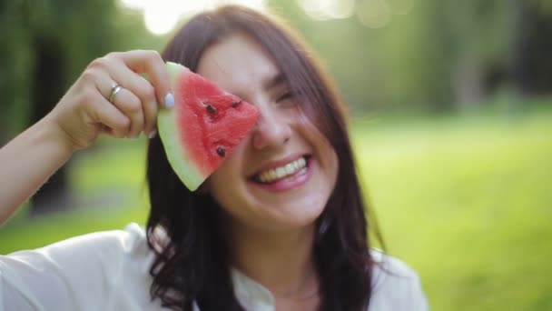 Schattig mooi brunette meisje met speelse humeur grimaces en lacht om de camera, eten plakje watermeloen op de achtergrond van gras park, close-up uitzicht. Gezicht van mooie en vrolijke jonge vrouw met fruit. — Stockvideo
