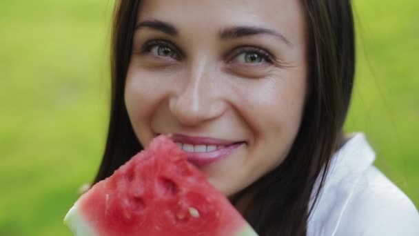 Detailní pohled na krásné evropské brunetky mladé ženy s laskavýma očima a těší jíst meloun na pozadí trávy v městském parku. Koncept volného času a aktivního života od atraktivní dívky — Stock video