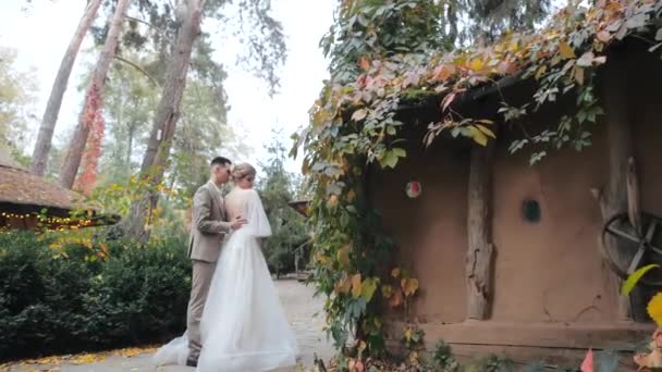 Μέση άποψη του νεαρού γαμπρού με κοστούμι αγκαλιάζει την αγαπημένη του νύφη με πολυτελές φόρεμα δίπλα σε άνετα εξοχικά σπίτια, καλυμμένα με φύλλα και βρίσκονται στο δάσος. Ευρωπαϊκή γάμο ζευγάρι βόλτες στο πάρκο φθινόπωρο. — Αρχείο Βίντεο