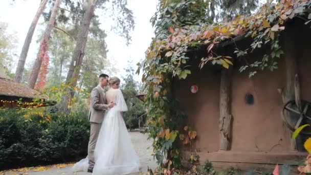 Vista media del joven novio en traje abrazando a su amada novia en lujoso vestido al lado de acogedoras casas de campo, cubiertas de hojas y ubicadas en el bosque. Pareja de boda europea camina en otoño parque . — Vídeos de Stock