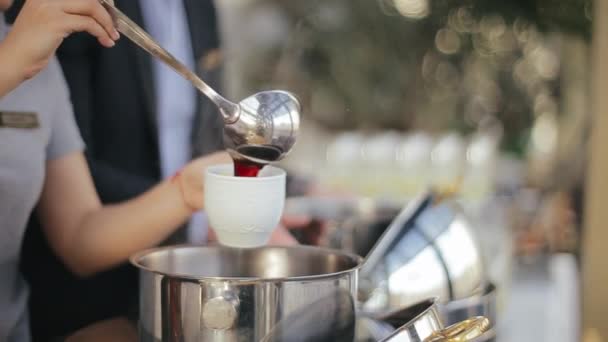 Kvinnlig servitör med ett rött armband på handen häller ett rött slag från en kastrull med en slev i en vit kopp för gäster på en bröllopsfest i festsalen. Personalen häller upp en varm alkoholdryck. — Stockvideo