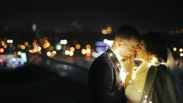 Caucásico joven novio en traje y novia en un abrigo de piel, envuelto en amarillo guirnalda festiva beso en balcón fuera respetable restaurante en el centro de la ciudad en el fondo de las linternas en la noche . — Vídeo de stock