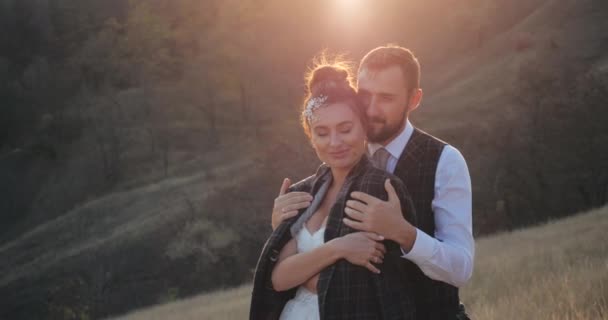 Close-up shot of cute married millennial couple hugging in a deserted spikelet field on warm sunny day. Carring groom wraps his beautiful bride in elegant white wedding dress with jacket. Rest outside — Stock Video