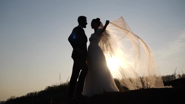 Vista cercana de las siluetas de la feliz pareja de boda de pie en la colina sobre el telón de fondo de una hermosa puesta de sol. Bonita novia en vestido con gracia baja el velo y mira al novio barbudo . — Vídeos de Stock