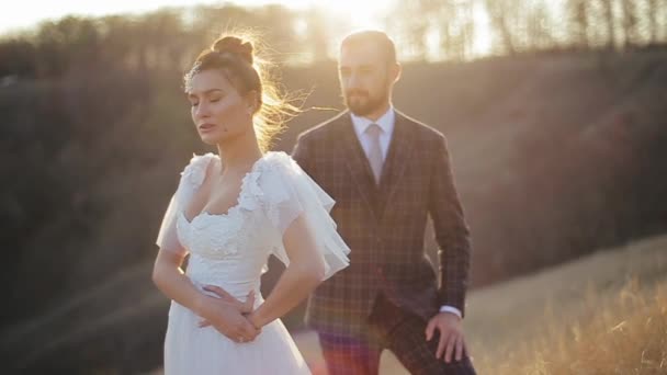 Jolie mariée dans une belle robe de mariée blanche sur le terrain regarde attentivement sur le côté, et son marié dans un costume se frotte le dos, gros plan, se concentrant sur une femme. Couple de mariage dehors dans la nature. — Video