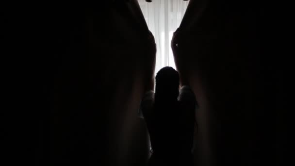 Achterfoto van de Kaukasische bruid in doorzichtig turquoise kamerjas met lang haar, die de gordijnen bij het raam opent om een donkere kamer in de ochtend te verlichten. Meisje trekt zwarte gordijnen terug. — Stockvideo